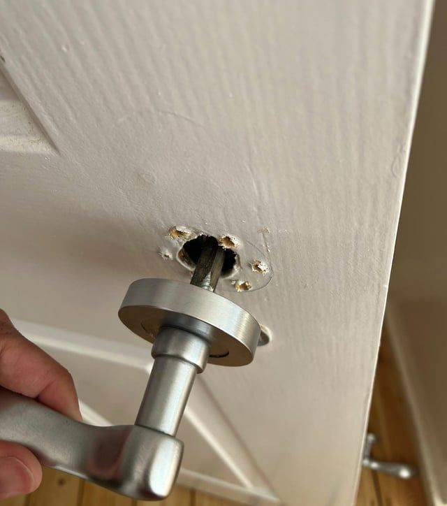 Hånd som monterer et nytt, sølvfarget dørhåndtak på en hvit dør med synlig hull.